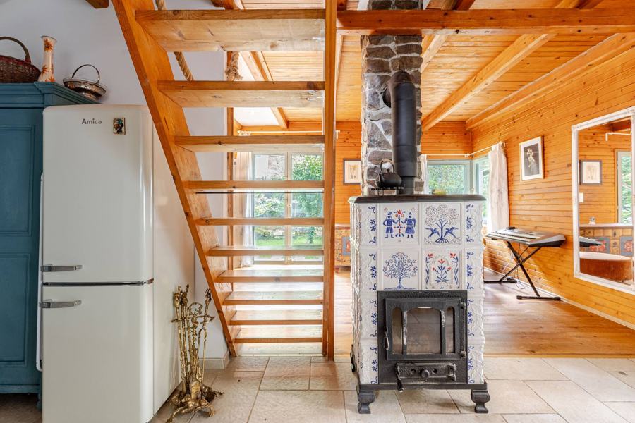 Bílá kachlová kamna s modrými ornamenty u dřevěného schodiště, vedle zelená skříň a retro lednice